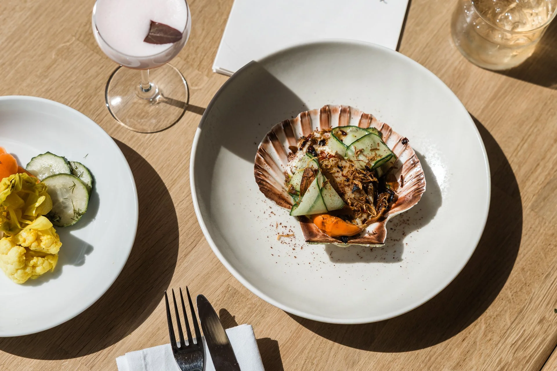 Hand-dived scallop served in its shell beside house pickles and a blush cocktail, on a sunlit oak table